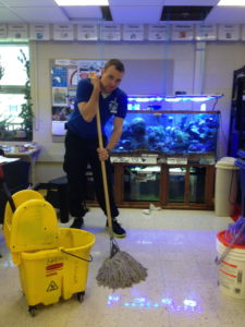 A male teacher is standing in front of an aquarium with a mop looking tired. The teacher was working overtime to clean up spilled water caused by a student mistake. A float switch on the water reservoir solved this problem.